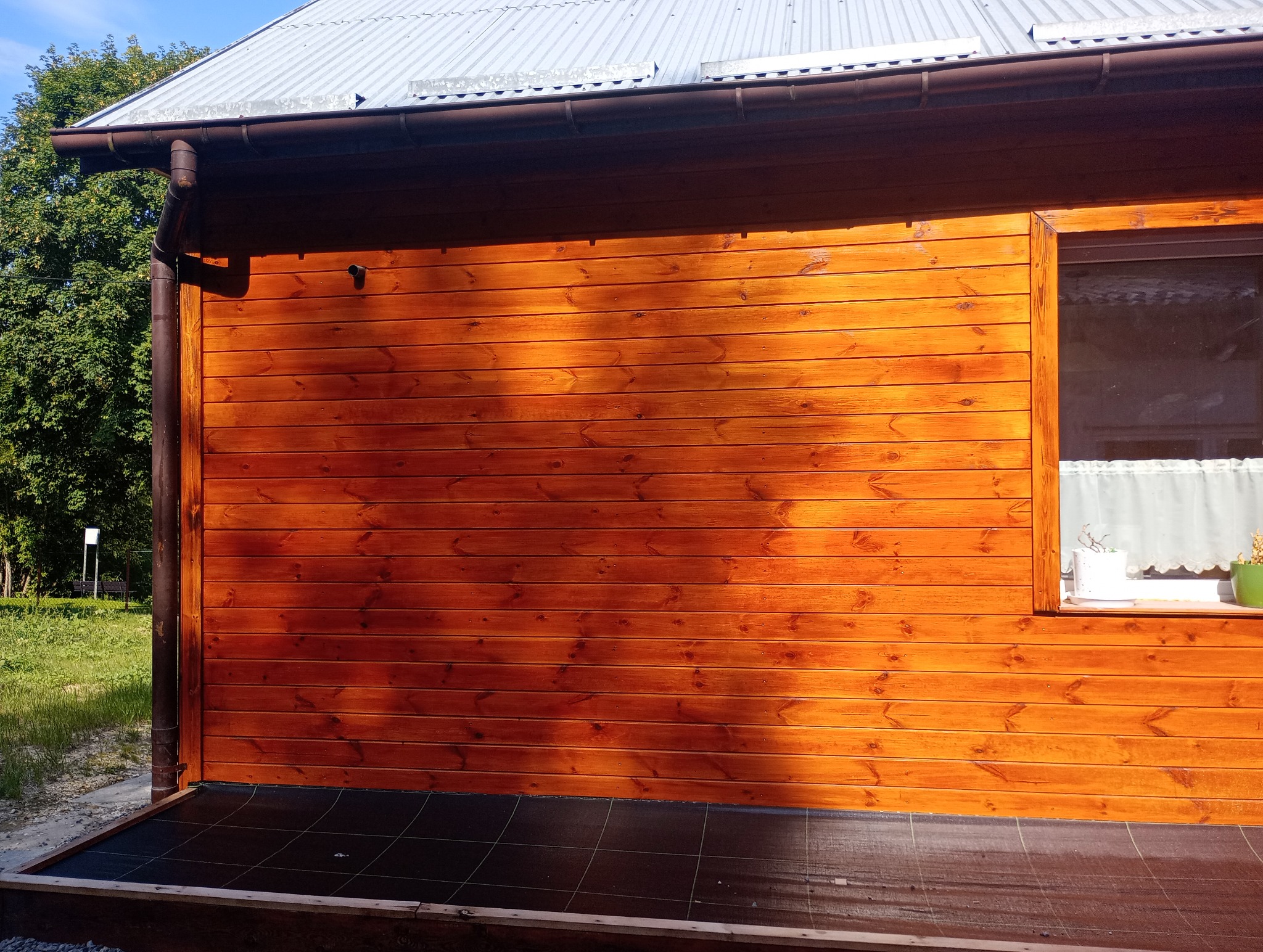 Ściana domu obita lakierowanym drewnem w odcieniach brązu, z widoczną rynną, oknem z firanką i kwiatami w doniczkach na parapecie, oraz fragmentem tarasu wyłożonego ciemnymi płytkami.