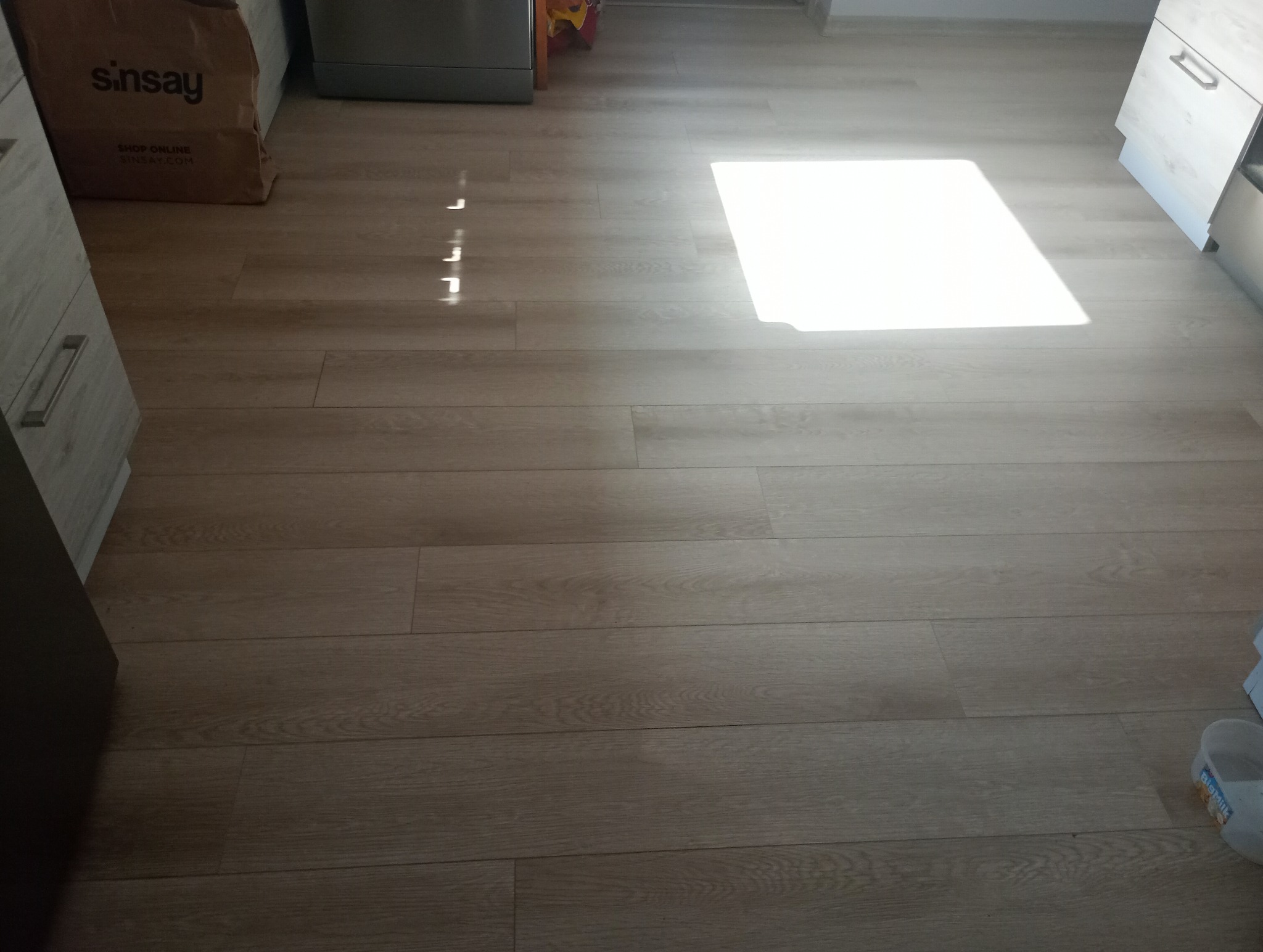 Jasne panele podłogowe ułożone w kuchni, z widocznym fragmentem szafki i torbą na zakupy w rogu, oświetlone silnym, naturalnym światłem słonecznym.