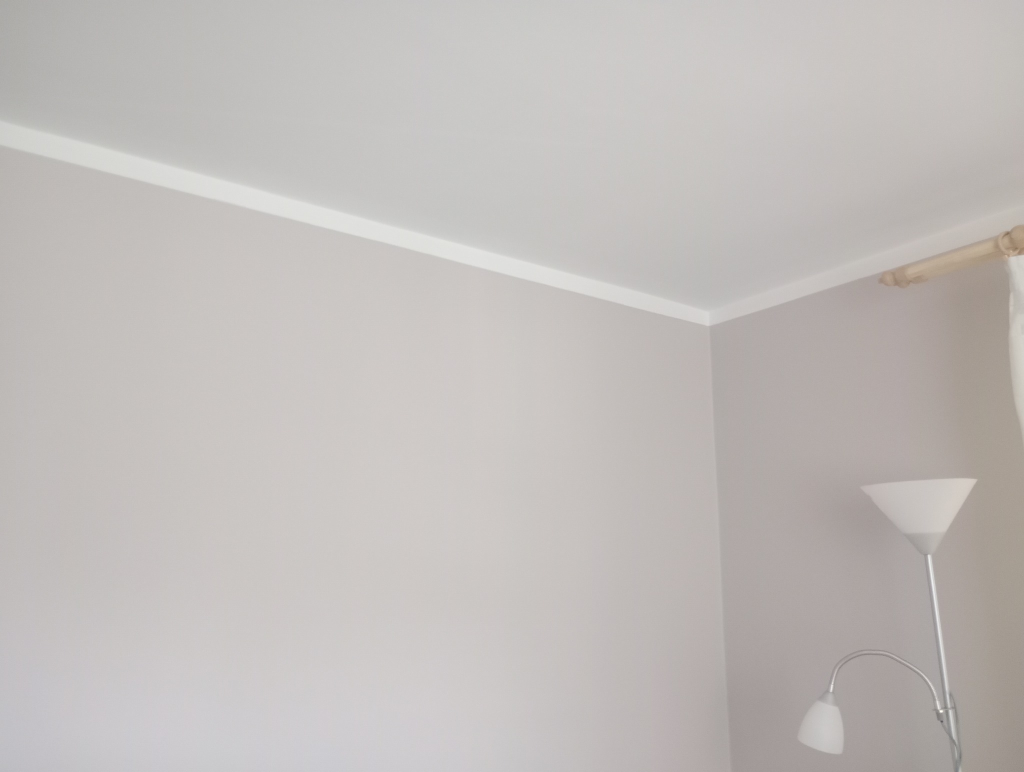 Jasnoszara ściana z białym, dekoracyjnym pasem pod sufitem, widoczny fragment karnisza i lampa podłogowa.
