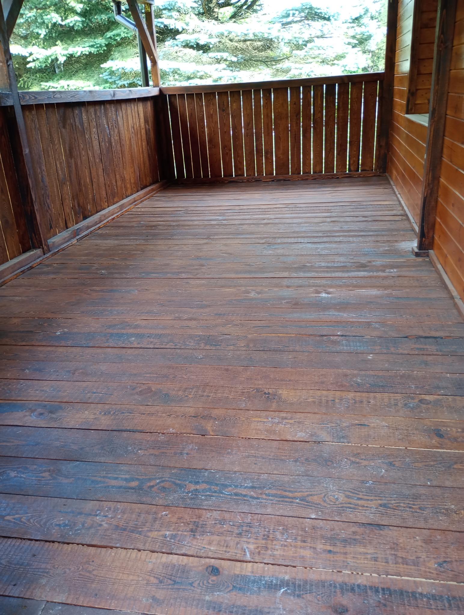 Drewniany taras z balustradą z pionowych desek, widok na zieleń drzew w tle.