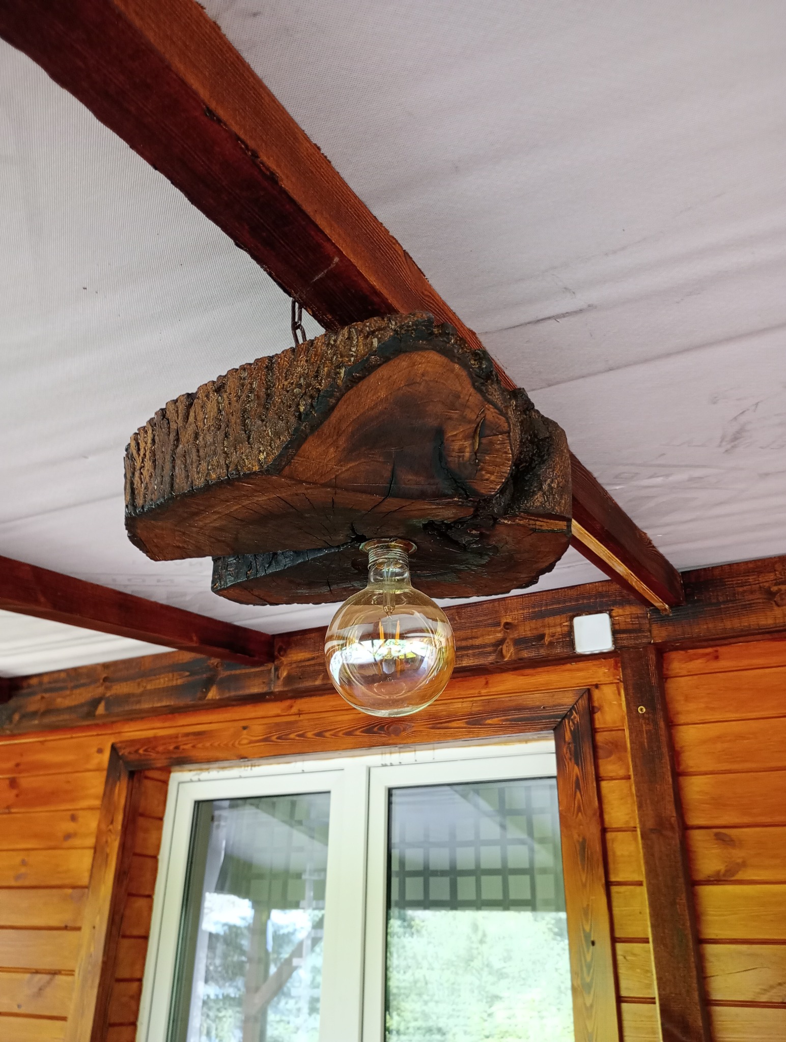 Drewniana lampa sufitowa wykonana z kawałka pnia drzewa, zwisająca nad oknem w drewnianej altanie.