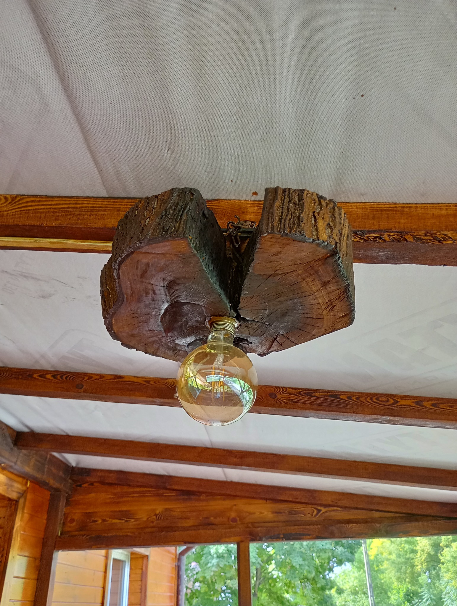 Nietypowa lampa sufitowa z drewnianego bala, zawieszona na łańcuchu, z żarówką w stylu retro, widoczna na tle drewnianej konstrukcji werandy i białego sufitu.