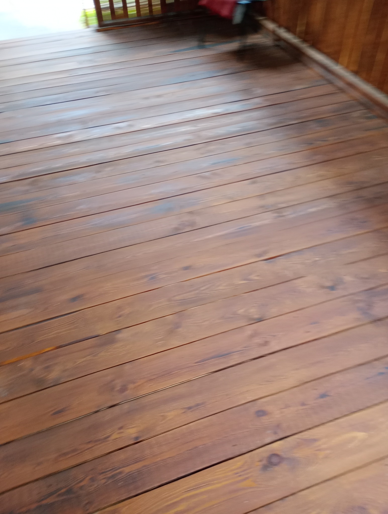 Drewniana podłoga tarasu z widocznymi słojami i brązową bejcą, świeżo po renowacji, z fragmentem balustrady w tle.