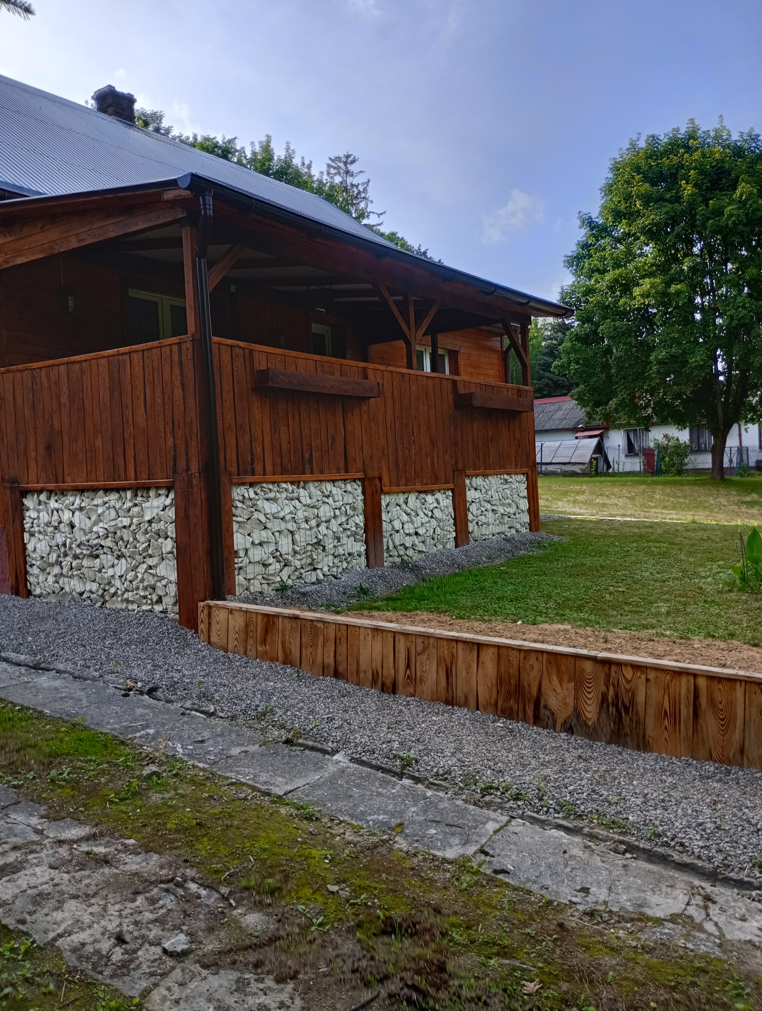 Drewniany dom z zadaszonym tarasem, ściany ozdobione gabionami z białego kamienia, drewniana palisada i wysypany żwir na podwórku.