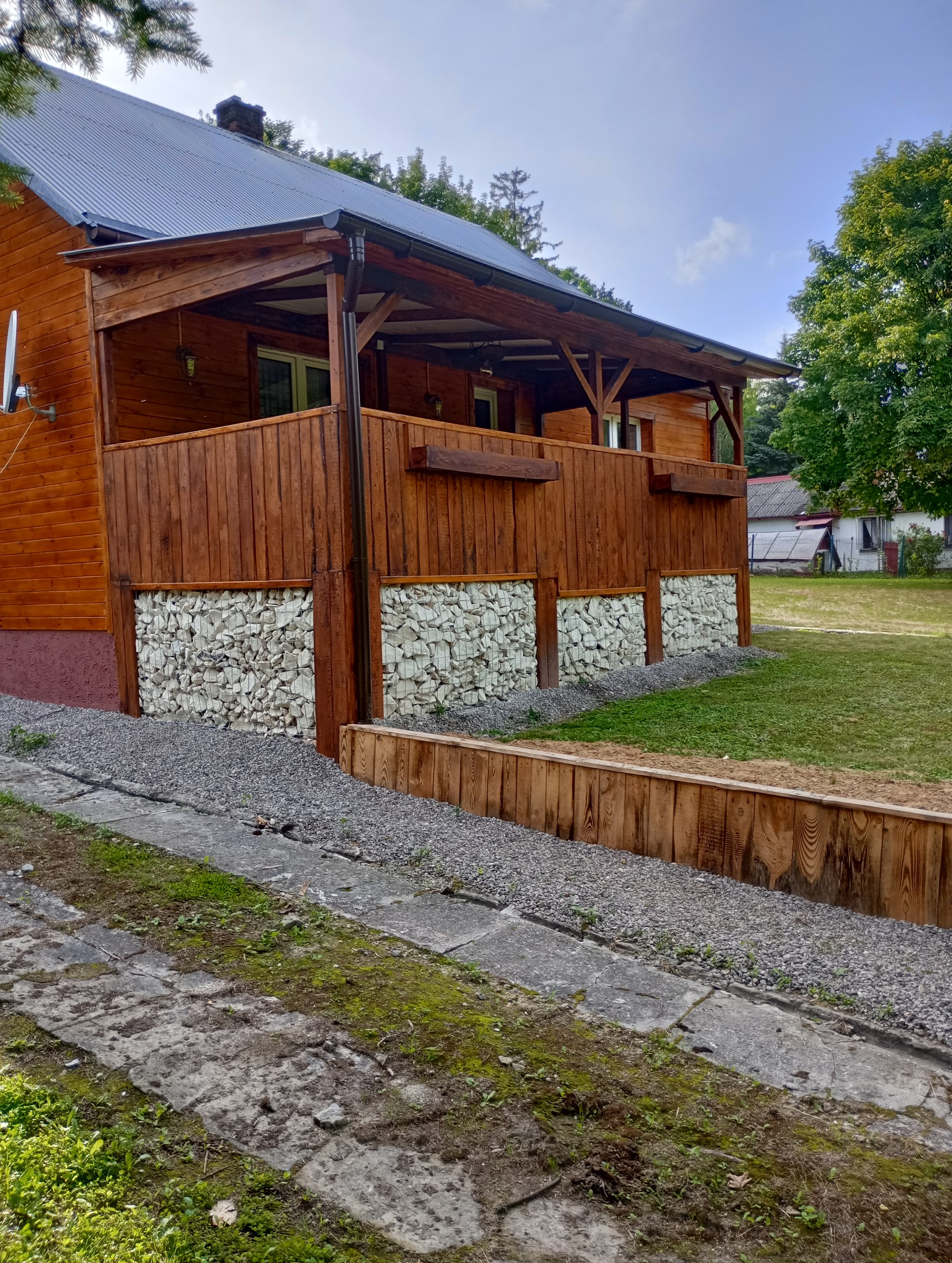 Drewniany dom z tarasem, podmurówka z gabionów wypełnionych białymi kamieniami, widok z zewnątrz na tle zieleni.
