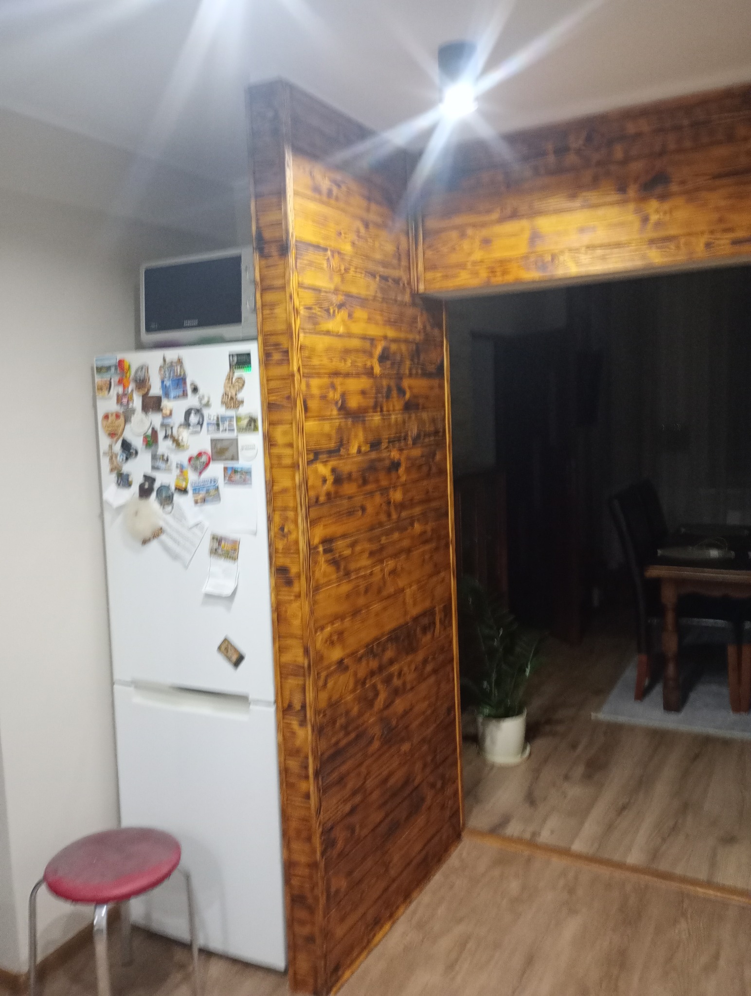 Wykończenie otworu drzwiowego w kuchni z drewnianą ramą o wyraźnym usłojeniu, widoczna biała lodówka z magnesami i czerwony stołek na tle paneli podłogowych.