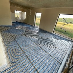 Bartłomiej Bombik - Niebieskie rury ogrzewania podłogowego ułożone spiralnie na czarnej siatce w surowym wnętrzu z dużymi oknami i stertą materiałów izolacyjnych w rogu.