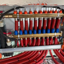 Bartłomiej Bombik - Rozdzielacz ogrzewania podłogowego z czerwonymi rurami w izolacji, widoczne zawory, odpowietrzniki i elementy montażowe w surowym otworze w ścianie.