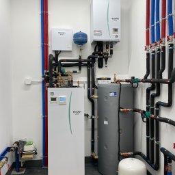 Bartłomiej Bombik - Nowoczesna kotłownia z dwoma zasobnikami, pompą ciepła Ecodan i rozbudowaną instalacją rur w kolorach czerwonym i niebieskim, z czarną izolacją, na tle białych ścian i ciemnej posadzki z widocznymi...
