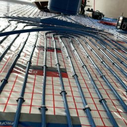 Bartłomiej Bombik - Montaż ogrzewania podłogowego: niebieskie rury układane na folii z siatką, widoczny pracownik w kombinezonie i narzędzia, duże okno w tle.