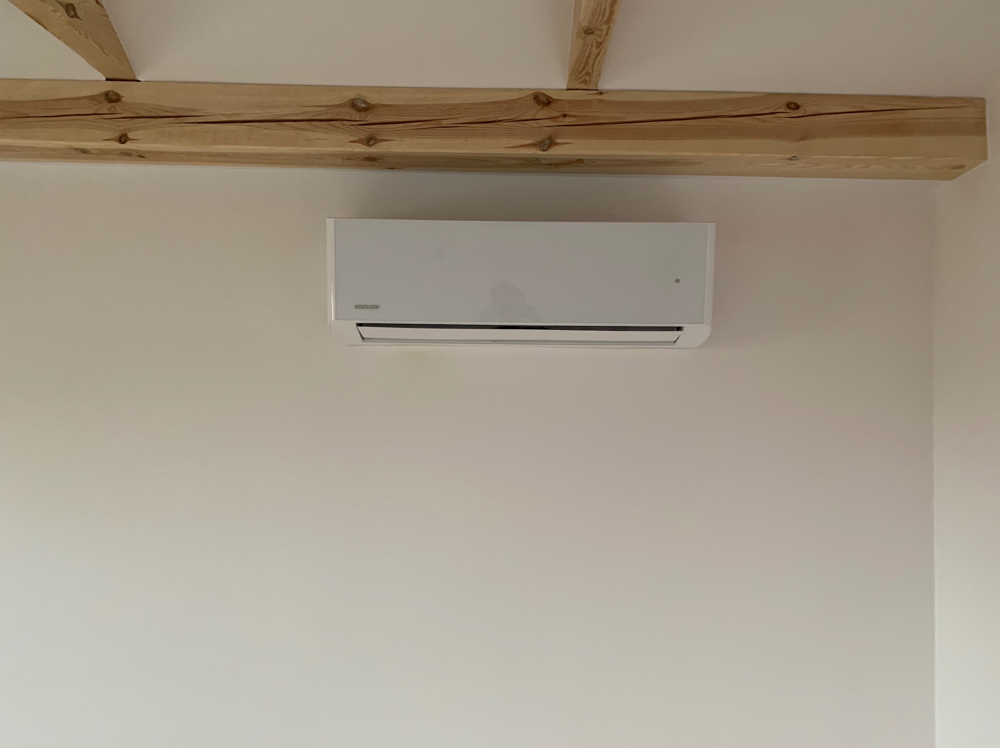 Biały klimatyzator ścienny zamontowany pod drewnianą belką stropową w jasnym pomieszczeniu.