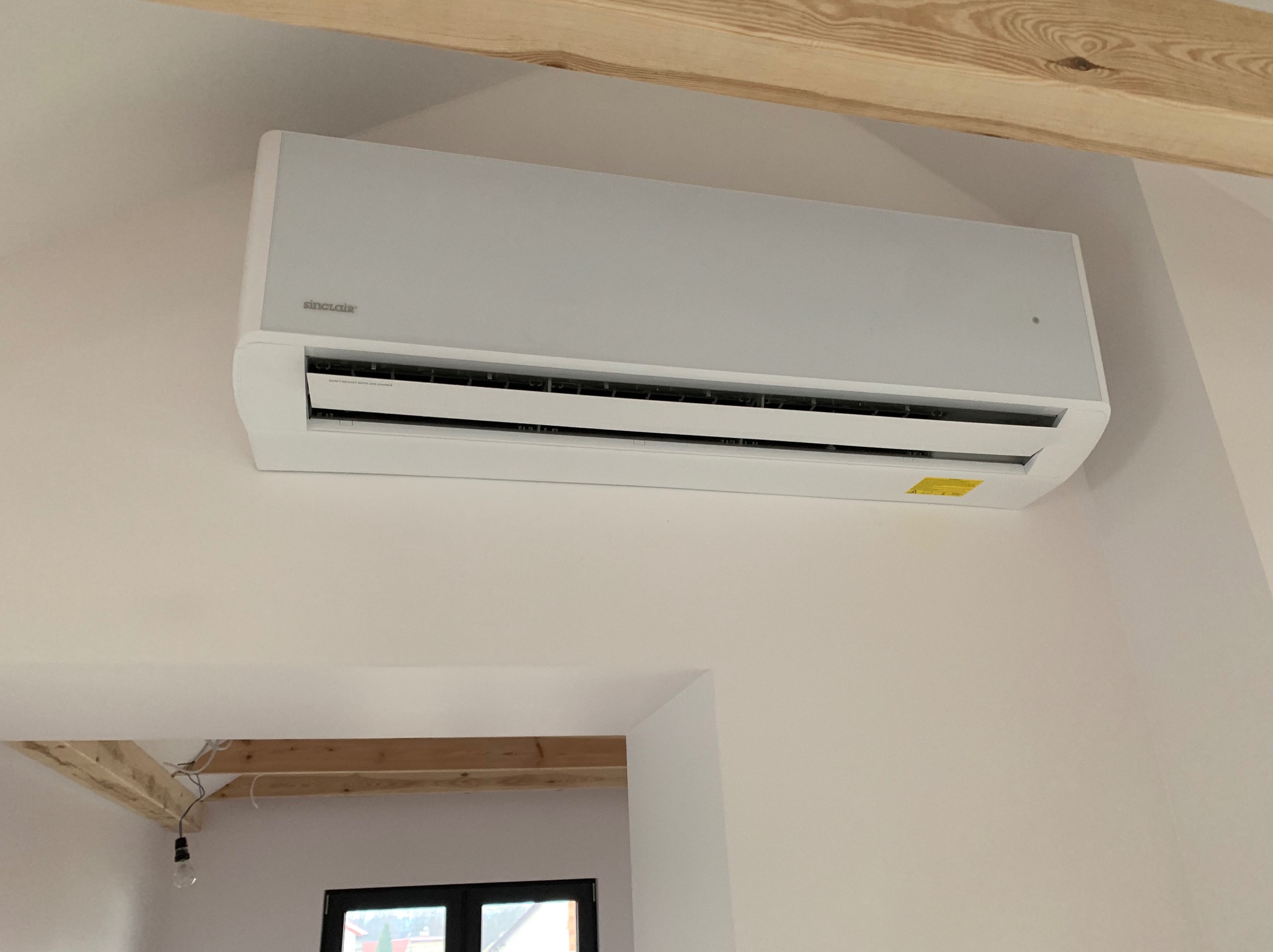 Biały klimatyzator marki Sinclair zamontowany na ścianie w nowym budynku, z widocznym drewnianym stropem i fragmentem wnętrza przez otwór drzwiowy.