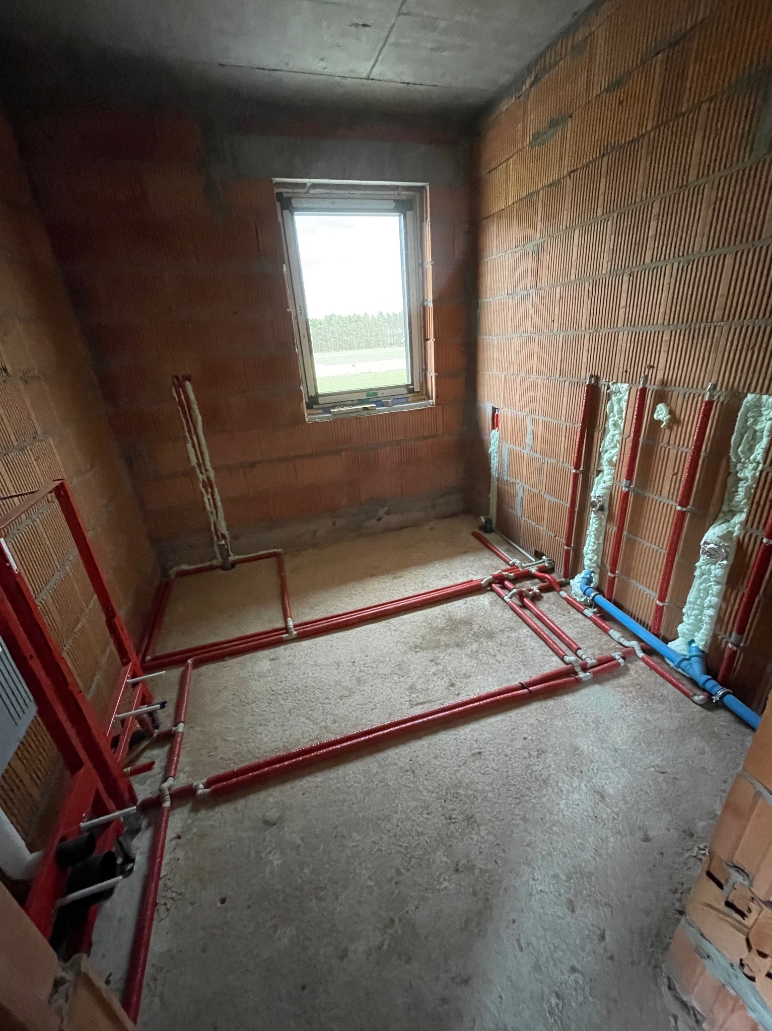 Surowy stan łazienki z widoczną instalacją wodną w kolorze czerwonym i niebieskim, pianką montażową uszczelnione przejścia przez ściany z cegły dziurawki, małe okno z widokiem na zieleń.