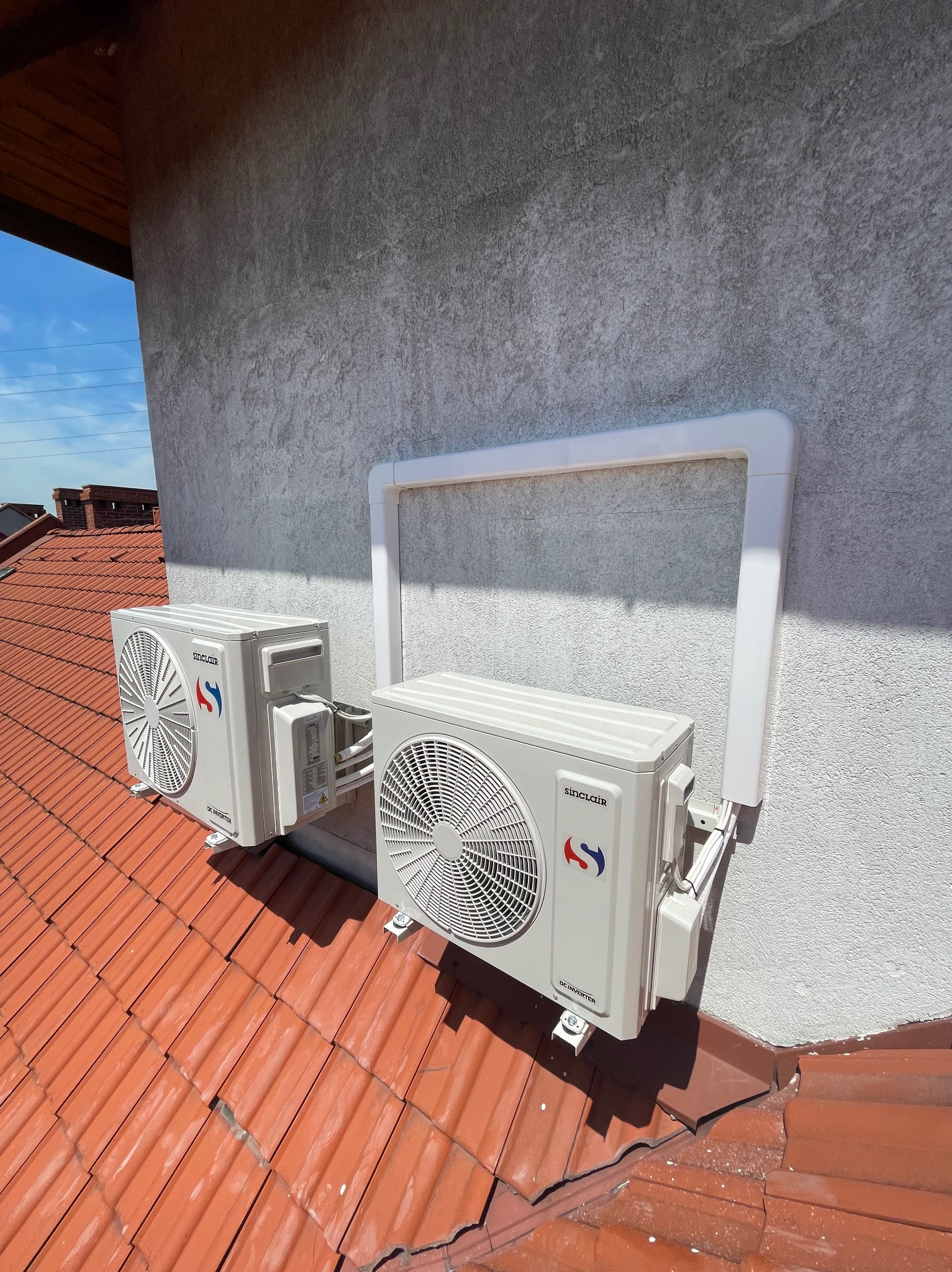 Dwa klimatyzatory Sinclair zamontowane na dachu z czerwonej dachówki, zabezpieczone białą ramą ochronną na elewacji budynku.