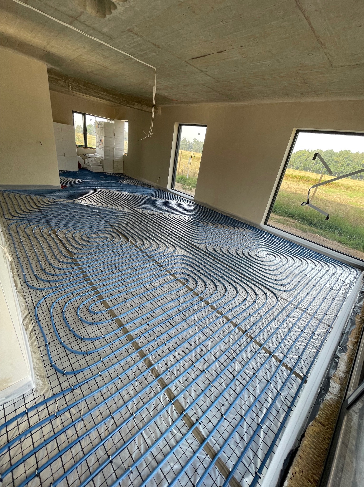 Niebieskie rury ogrzewania podłogowego ułożone spiralnie na czarnej siatce w surowym wnętrzu z dużymi oknami i stertą materiałów izolacyjnych w rogu.