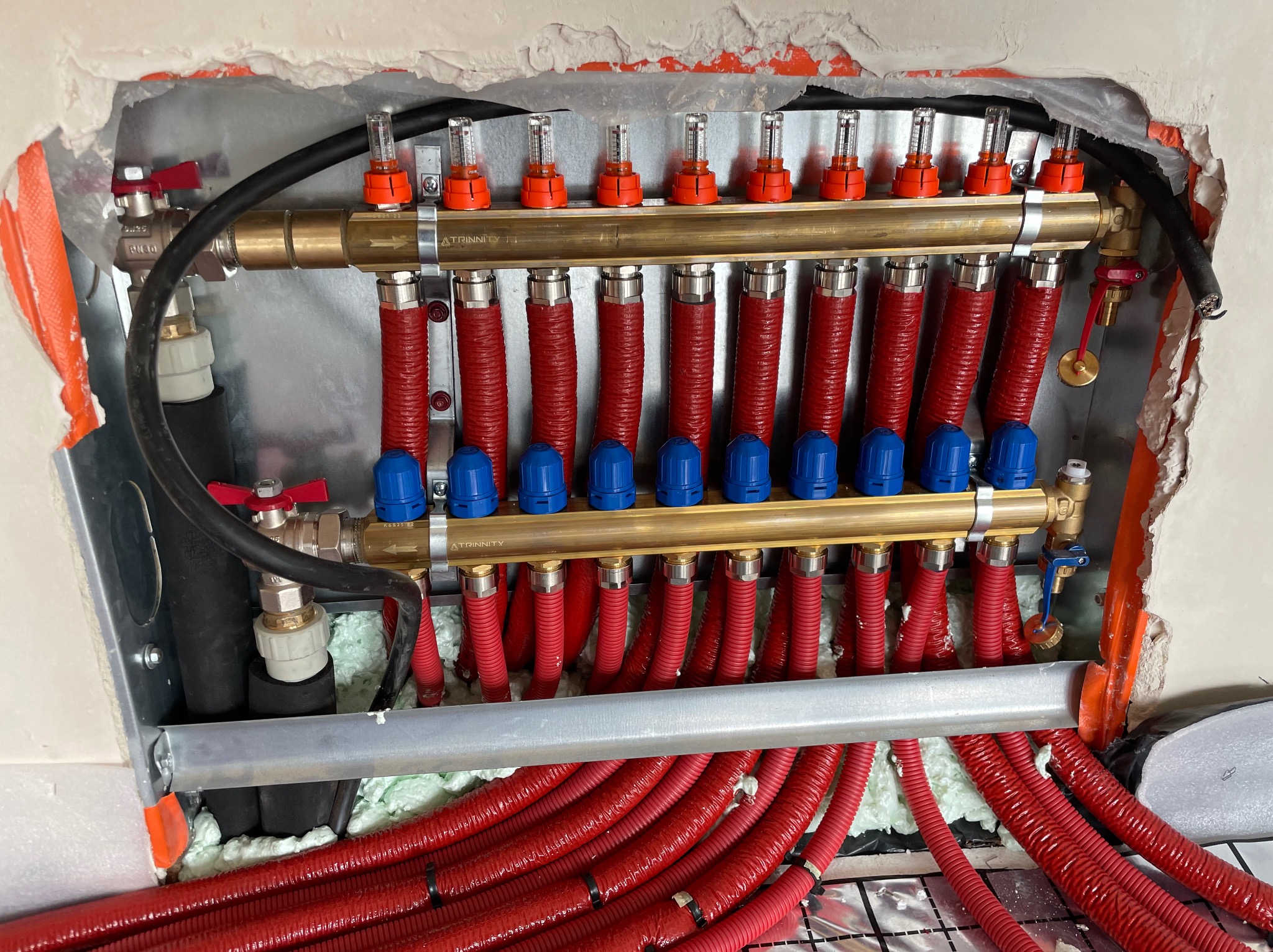 Rozdzielacz ogrzewania podłogowego z czerwonymi rurami w izolacji, widoczne zawory, odpowietrzniki i elementy montażowe w surowym otworze w ścianie.