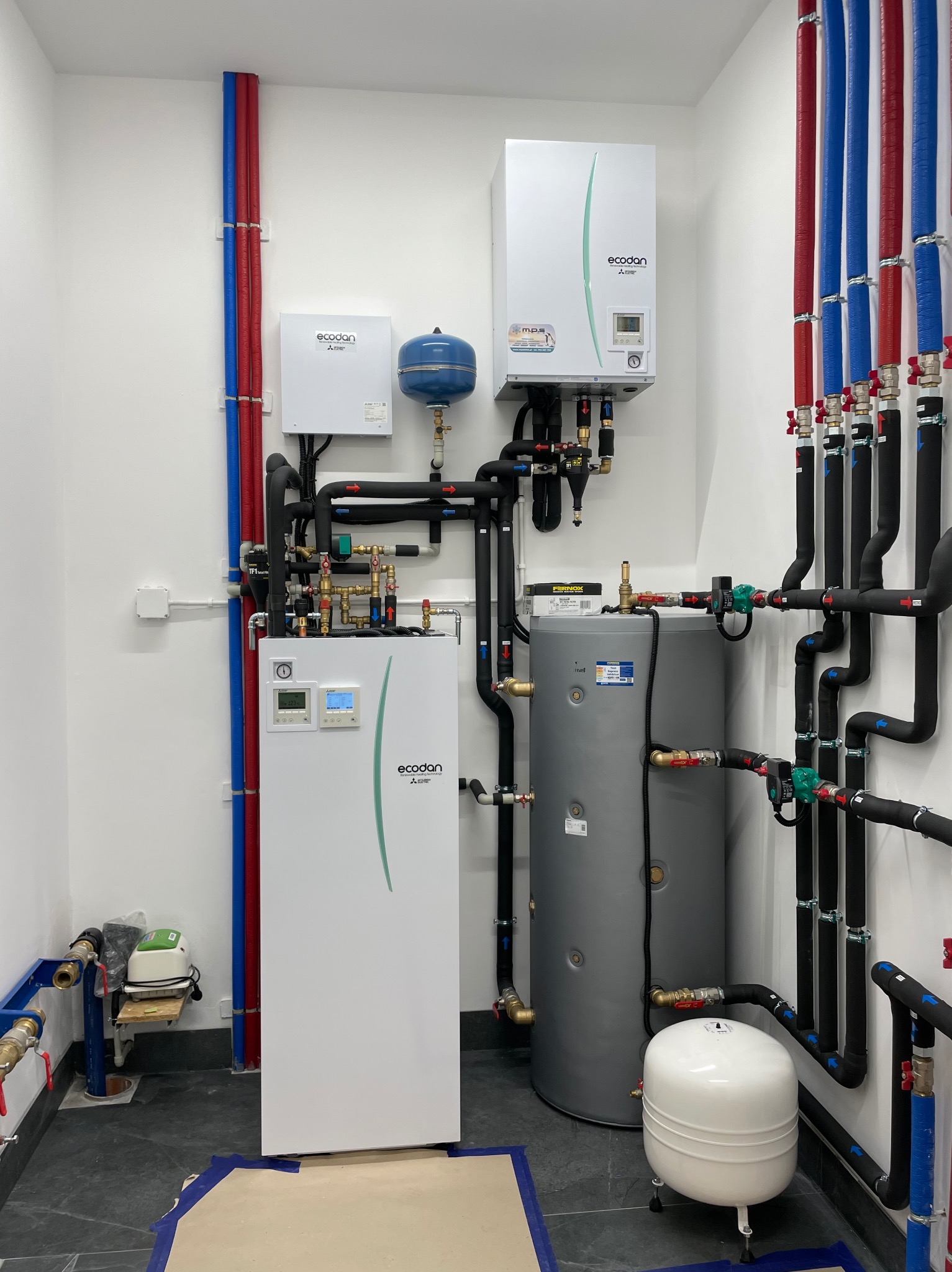 Nowoczesna kotłownia z dwoma zasobnikami, pompą ciepła Ecodan i rozbudowaną instalacją rur w kolorach czerwonym i niebieskim, z czarną izolacją, na tle białych ścian i ciemnej posadzki z widocznymi...