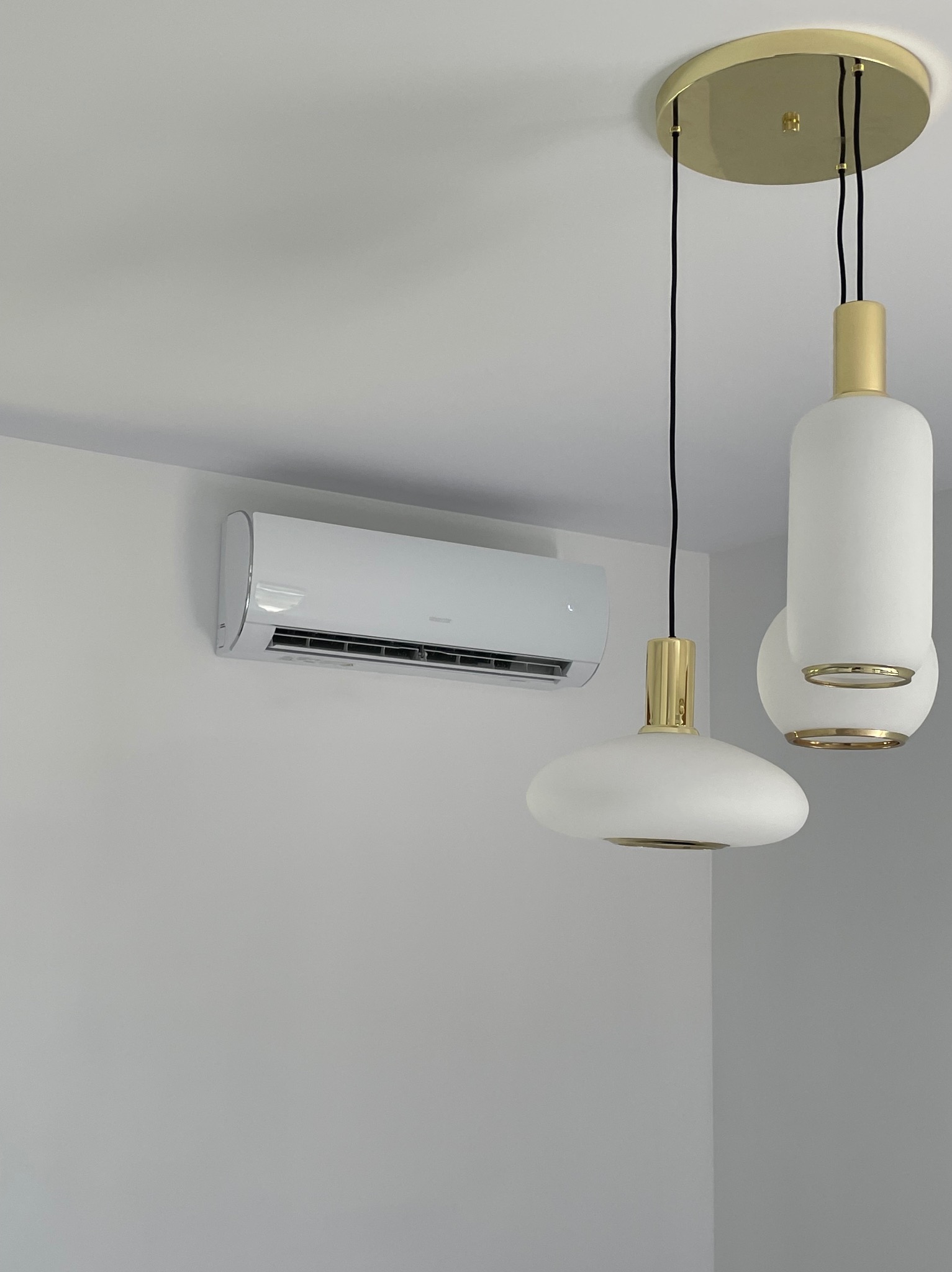 Biała jednostka klimatyzacji ściennej z widocznymi żaluzjami, zamontowana w rogu pomieszczenia, obok nowoczesnej, białej lampy wiszącej z elementami w kolorze złotym.