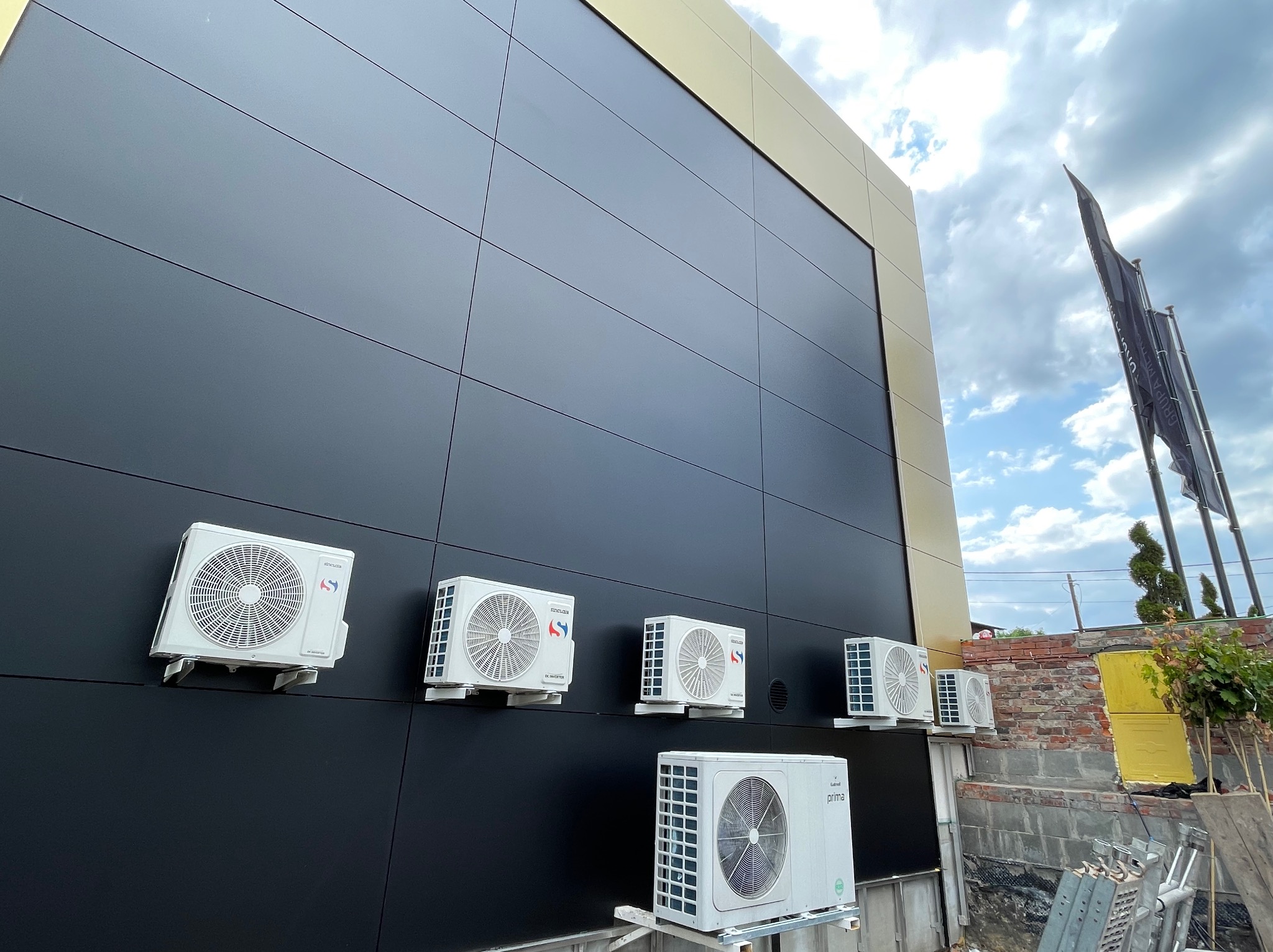 Elewacja budynku z sześcioma zamontowanymi jednostkami zewnętrznymi klimatyzacji, widoczne flagi firmowe i fragment rusztowania.