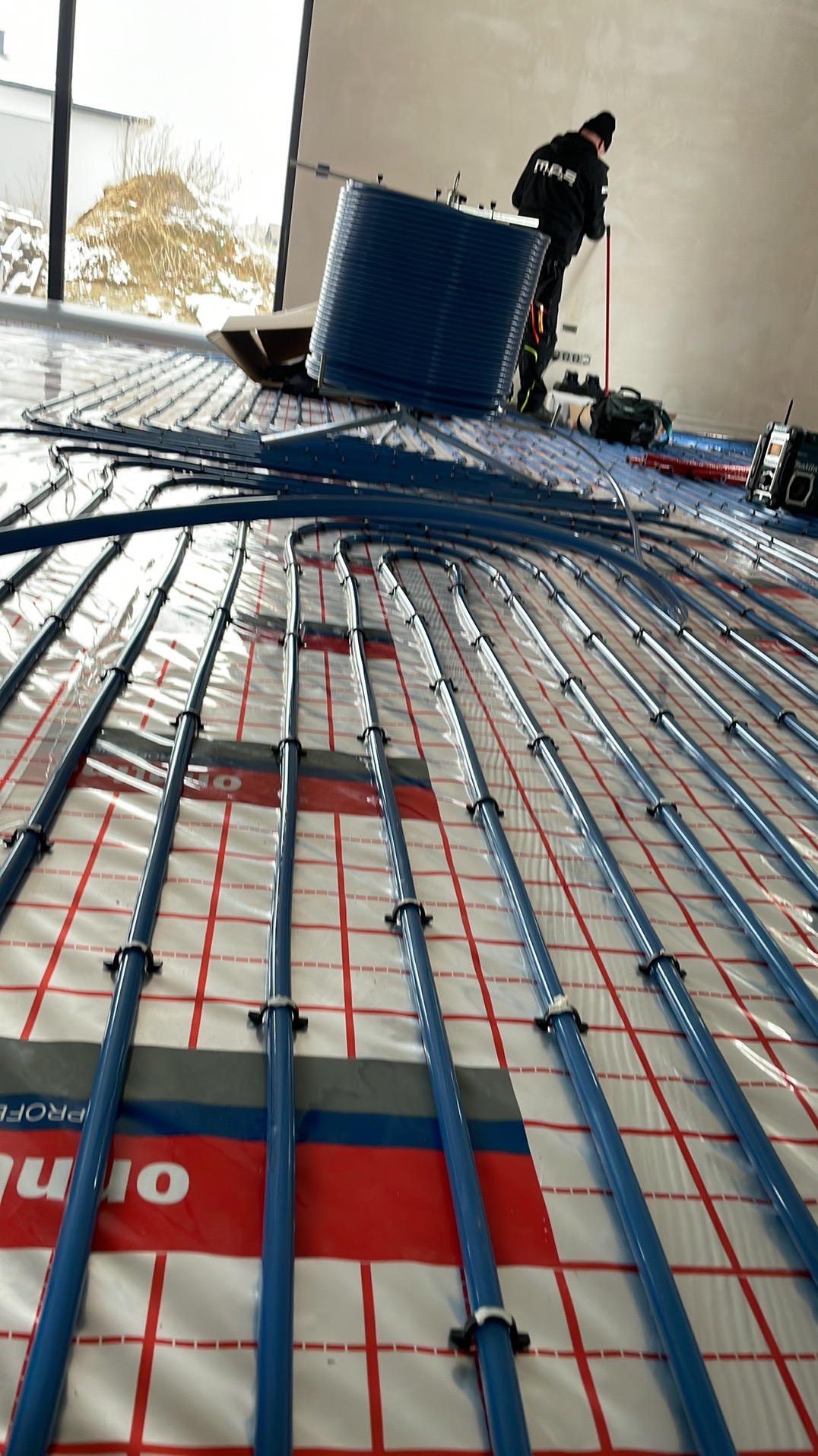 Montaż ogrzewania podłogowego: niebieskie rury układane na folii z siatką, widoczny pracownik w kombinezonie i narzędzia, duże okno w tle.