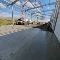 Wnętrze konstrukcji stalowej z betonową wylewką w trakcie realizacji; pracownicy wyrównują powierzchnię, w tle betoniarka z pompą do betonu.