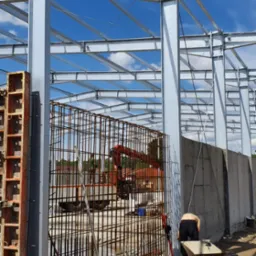 Konstrukcja żelbetowa w trakcie budowy: stalowe słupy, zbrojenie ścian, szalunki i pracownik z taczką na budowie w słoneczny dzień.