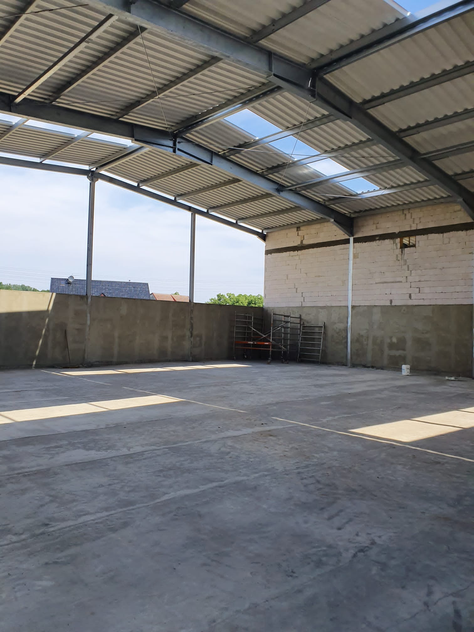 Wnętrze hali w stanie surowym: betonowa posadzka, ściany z betonu i bloczków, stalowa konstrukcja dachu z świetlikami, drabiny budowlane w tle.