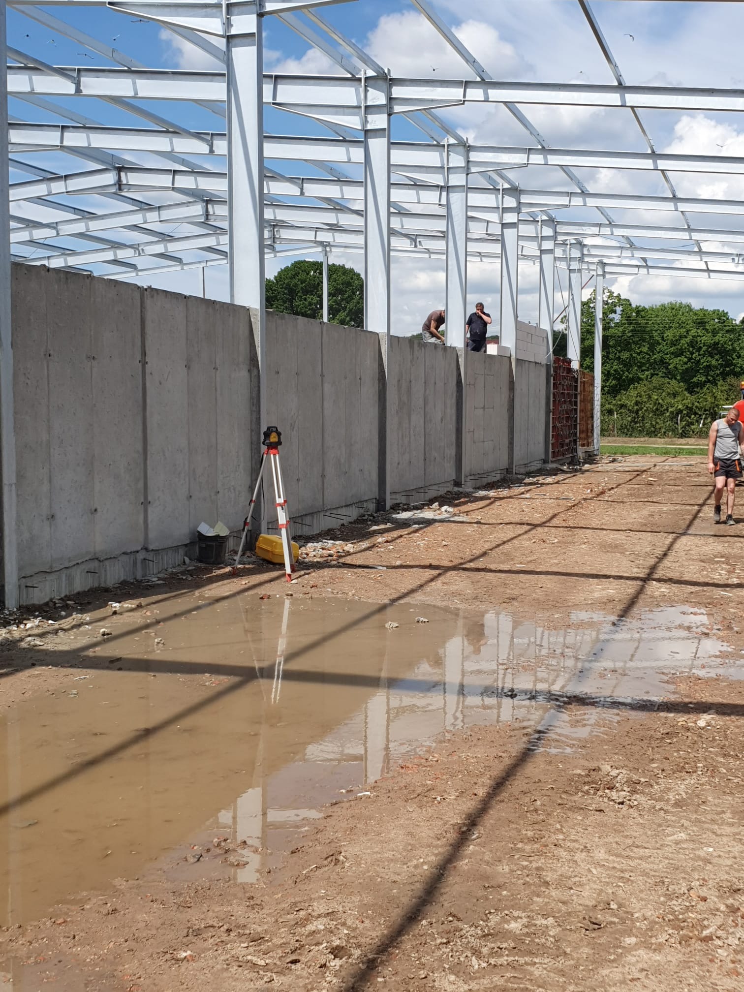 Budowa hali w Marszewie: wznoszenie ścian z prefabrykatów betonowych, widoczny stalowy szkielet konstrukcji dachu, poziomica laserowa i kałuża na placu budowy.