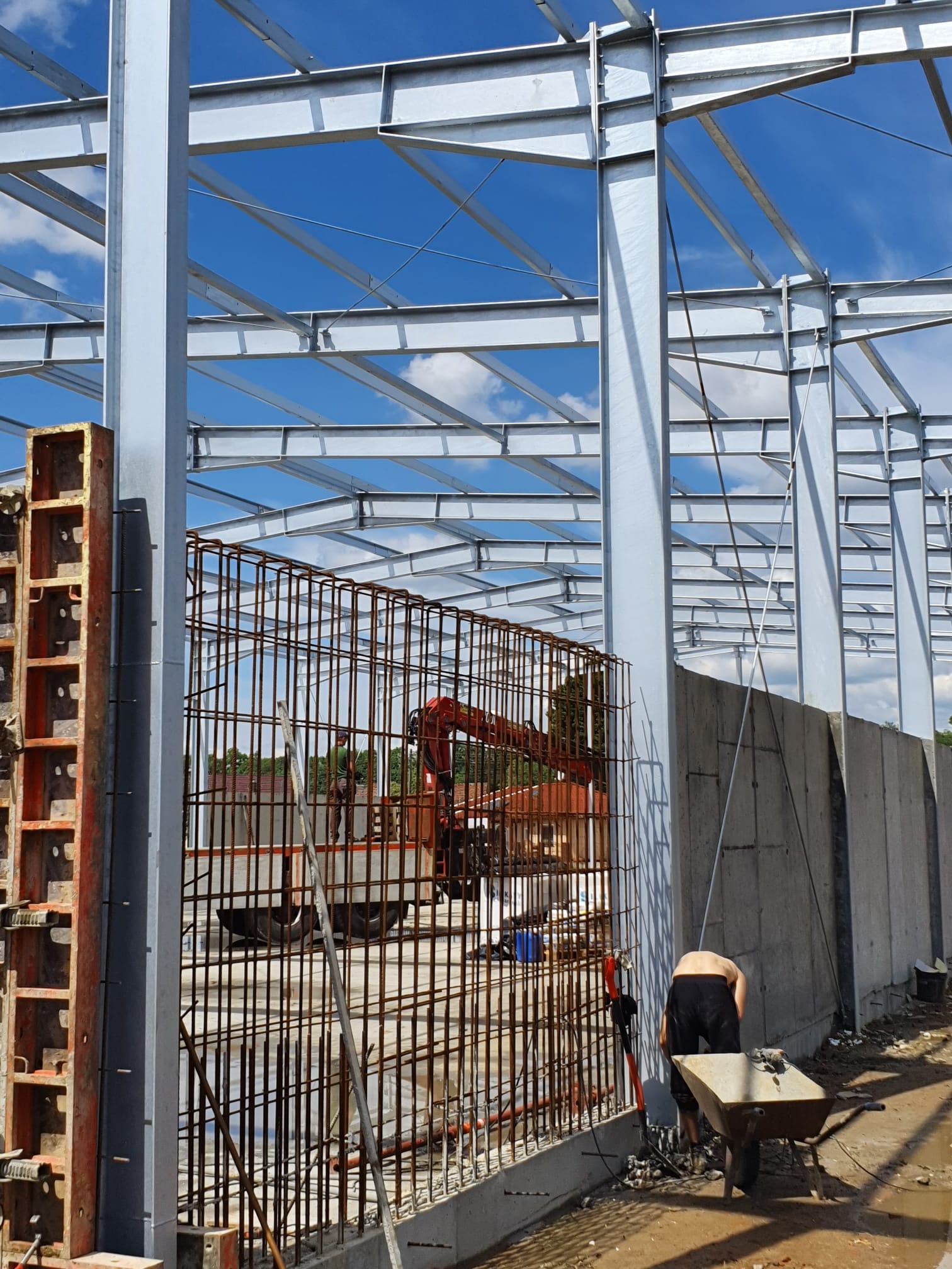 Konstrukcja żelbetowa w trakcie budowy: stalowe słupy, zbrojenie ścian, szalunki i pracownik z taczką na budowie w słoneczny dzień.