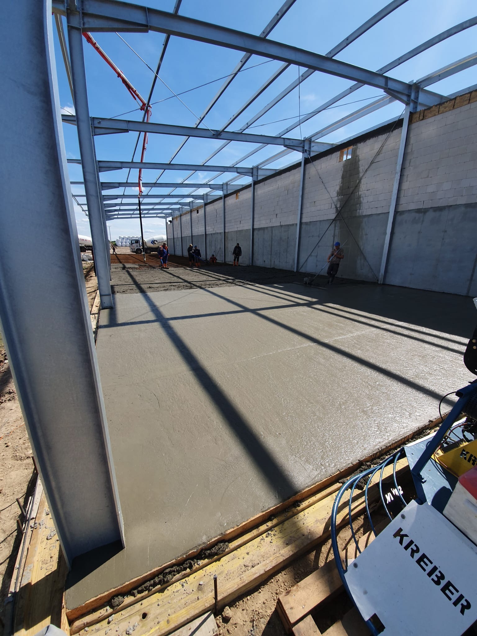 Wylewka betonu na hali produkcyjnej w trakcie realizacji, widoczna konstrukcja stalowa, pompa do betonu, ekipa budowlana i maszyna do zacierania betonu marki Kreber.