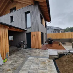 Nowoczesny dom z szarą elewacją i drewnianym wykończeniem, taras z drewnianą podłogą, fragment ułożonej kostki brukowej, widoczny drenaż liniowy i przygotowany teren pod ogród.