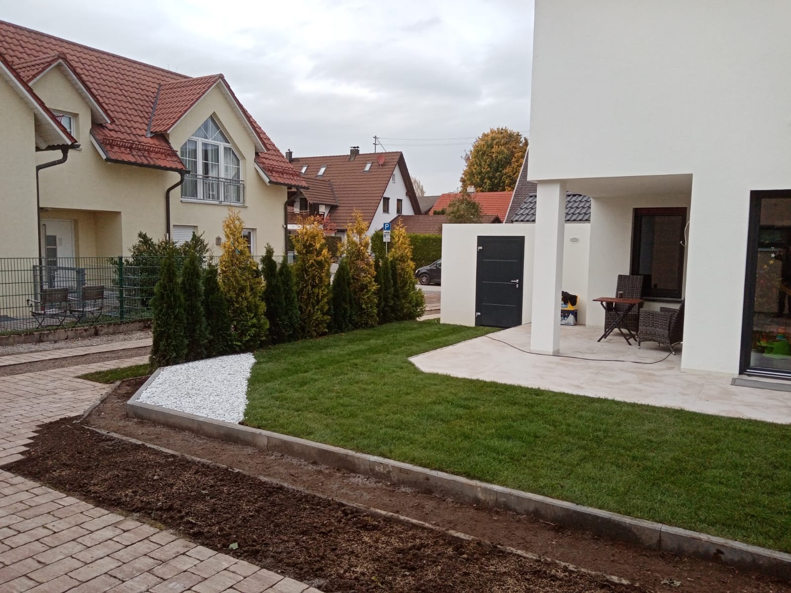 Nowo założony trawnik z ozdobnym białym grysem i rabatą przygotowaną pod nasadzenia, na tle nowoczesnego domu z tarasem i sąsiedniej zabudowy.