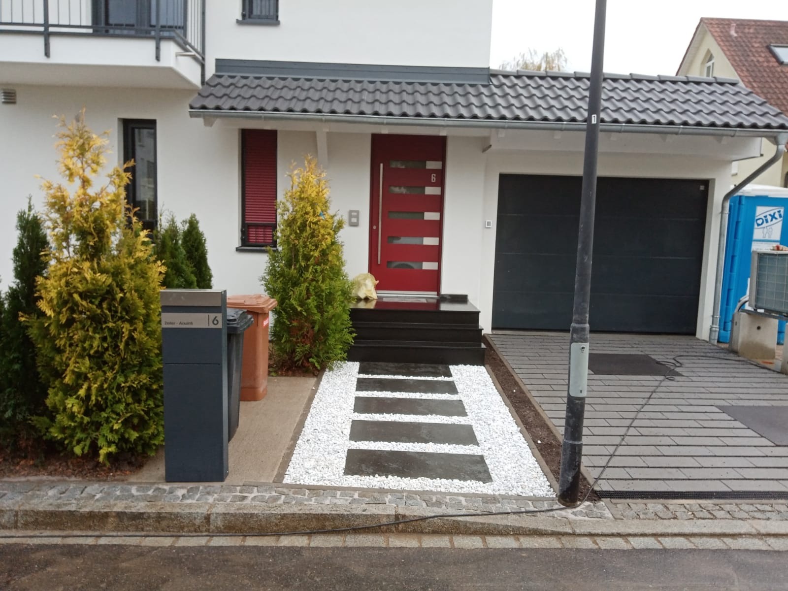 Nowoczesny dom z ciemnymi schodami wejściowymi i ścieżką z płyt otoczoną białym grysem, szara kostka brukowa na podjeździe, widoczne drzewka ozdobne i skrzynki na listy.