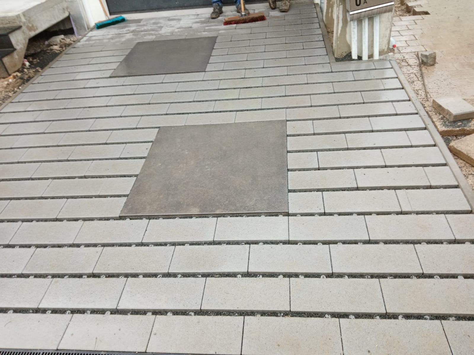 Nowa nawierzchnia z jasnoszarej kostki brukowej ułożonej w rzędach, z wkomponowanymi kwadratowymi płytami, częściowo zamieciona, widoczne buty robotników i szczotka.
