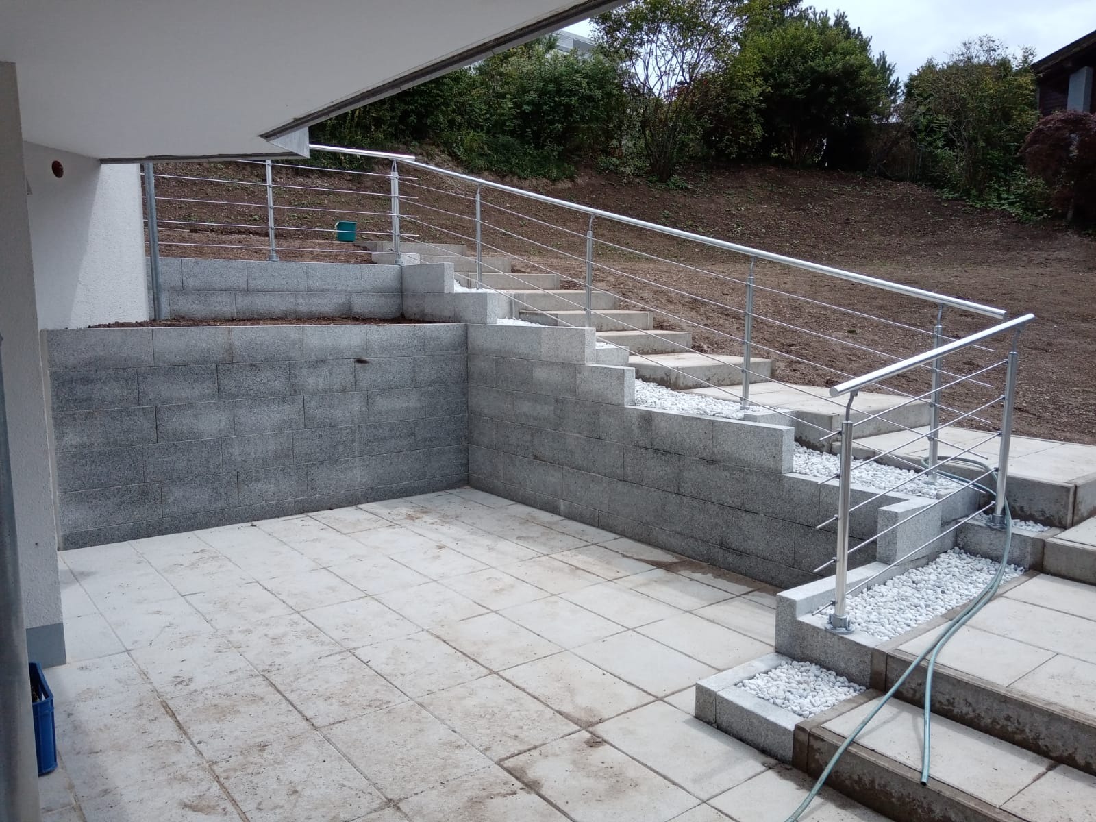 Taras wyłożony płytami z szarego kamienia, schody z bloków betonowych z metalową balustradą i białym grysem, widok na ogród.