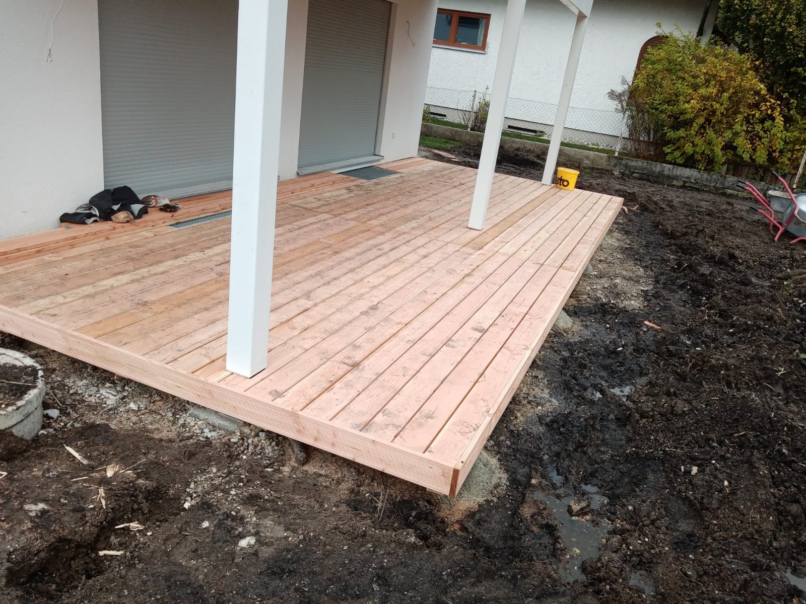 Drewniany taras w trakcie budowy, z białymi słupami podtrzymującymi zadaszenie, otoczony świeżo przekopaną ziemią i widocznymi narzędziami ogrodniczymi.