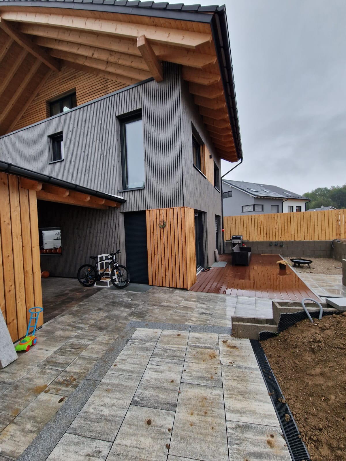 Nowoczesny dom z szarą elewacją i drewnianym wykończeniem, taras z drewnianą podłogą, fragment ułożonej kostki brukowej, widoczny drenaż liniowy i przygotowany teren pod ogród.