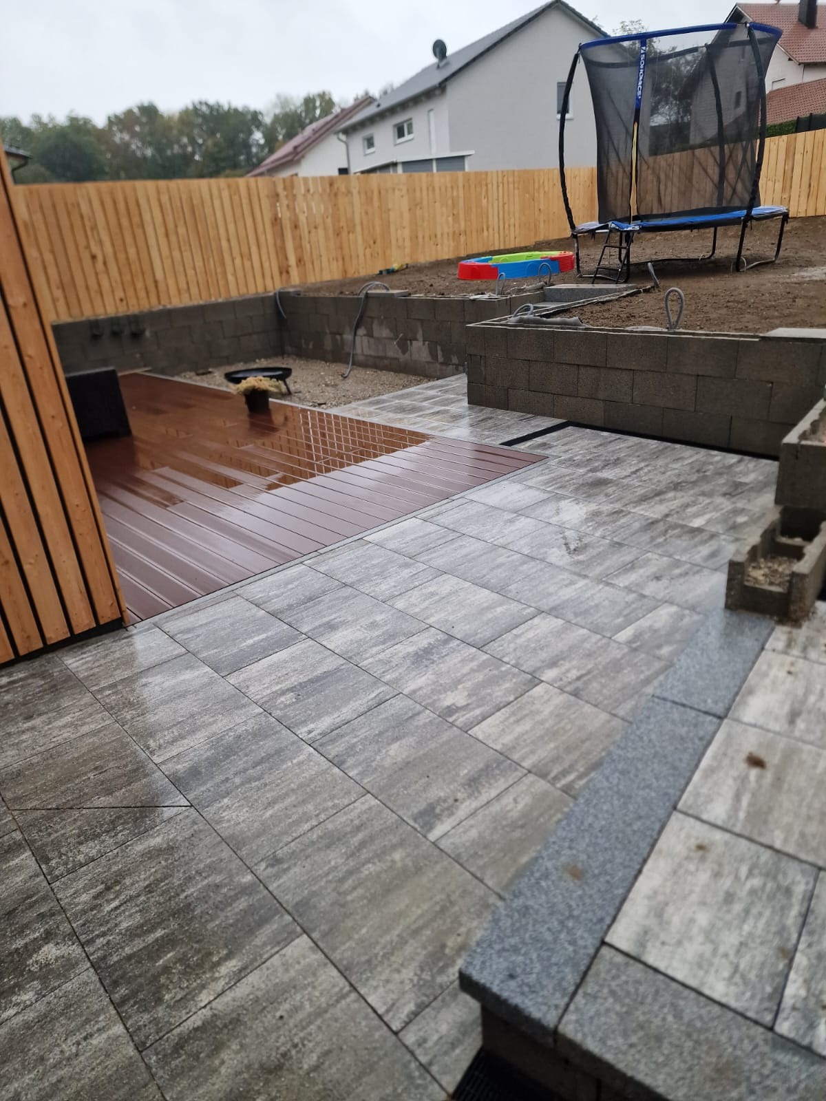 Ułożona szara kostka brukowa na posesji z drewnianym tarasem i ogrodzeniem w tle, widoczny trampolina i zabawki dla dzieci, pochmurna pogoda.