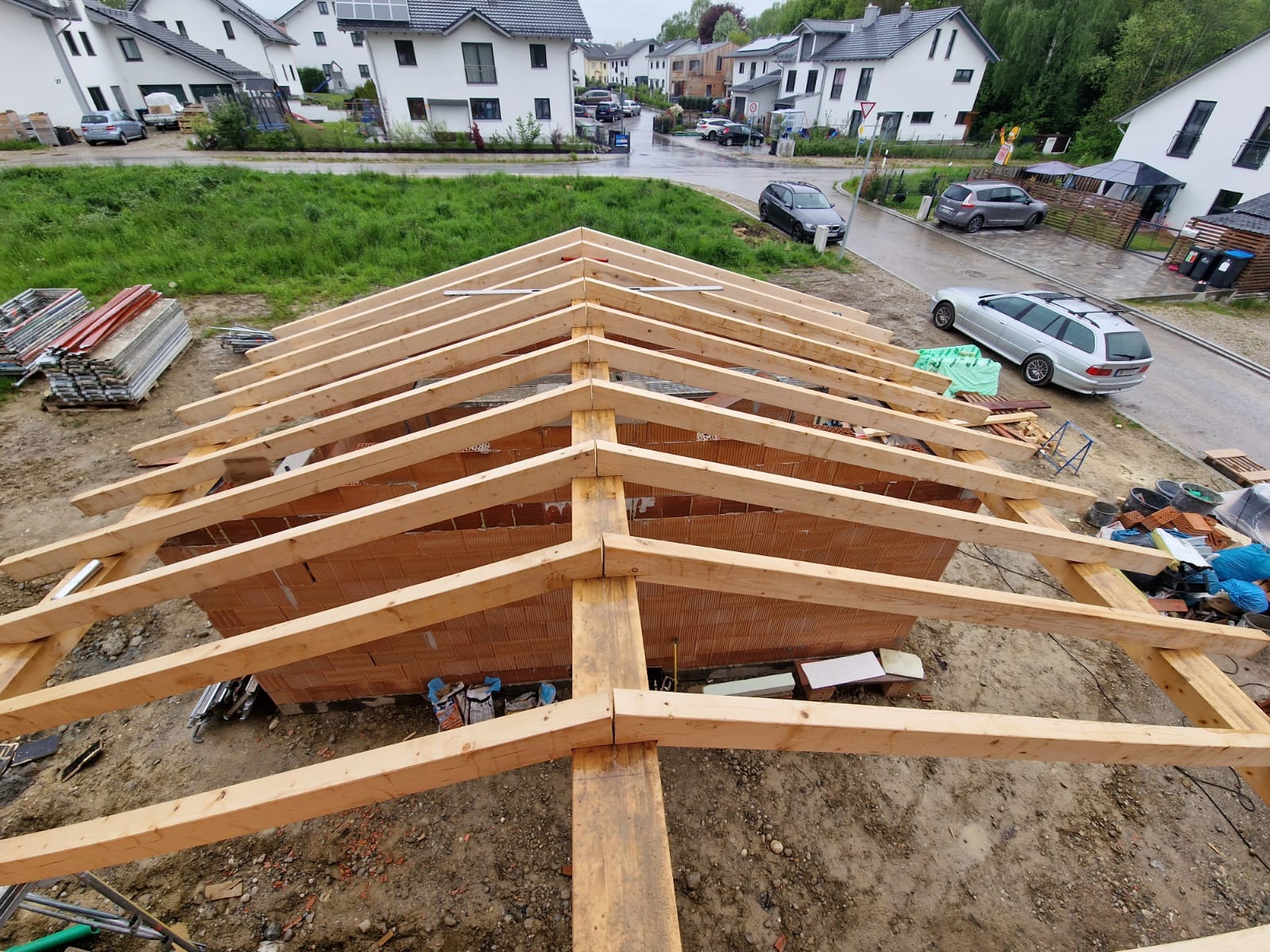 Konstrukcja drewniana dachu w trakcie budowy, widoczne cegły i elementy więźby dachowej, w tle zabudowa jednorodzinna i mokra ulica.