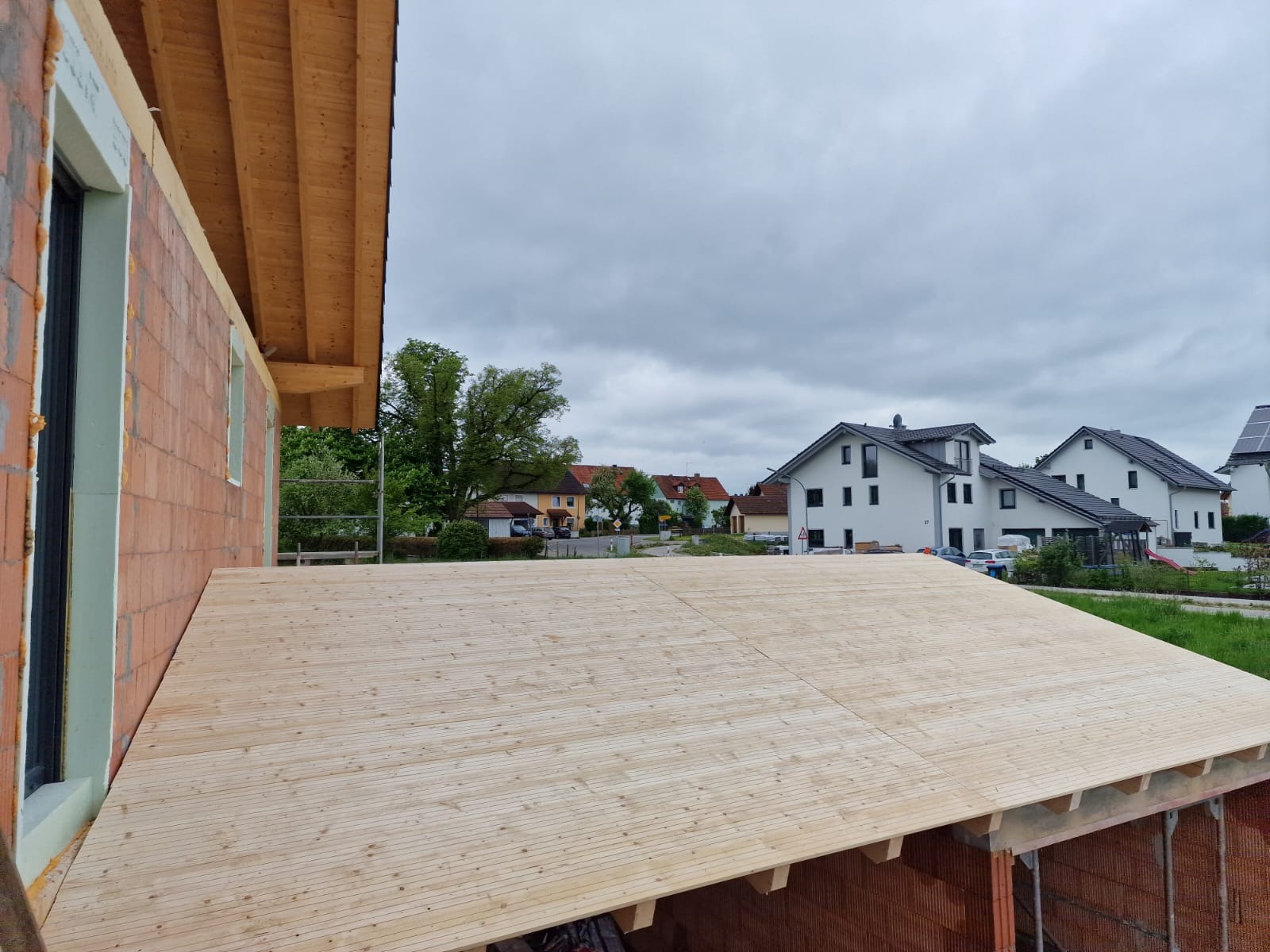 Surowa drewniana konstrukcja stropu domu jednorodzinnego w trakcie budowy, widok z góry na tle zabudowy mieszkalnej i pochmurnego nieba.