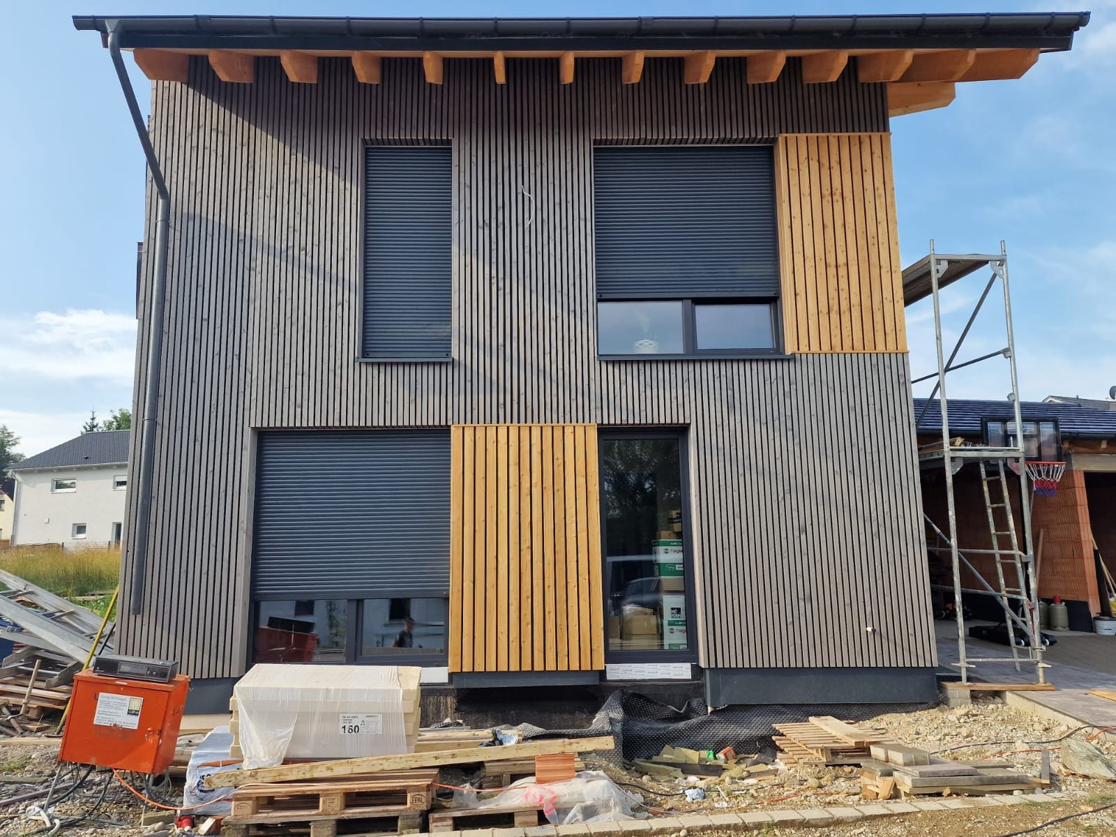 Nowoczesny dom w trakcie wykańczania elewacji z pionowych desek w dwóch odcieniach drewna, z częściowo zasłoniętymi roletami oknami i rusztowaniem obok.