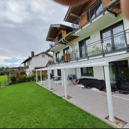 Aleksander Musioł - Nowoczesny dom z balkonami i zadaszonym tarasem wyłożonym szarą kostką, obok zadbany trawnik z zielonym ogrodzeniem.