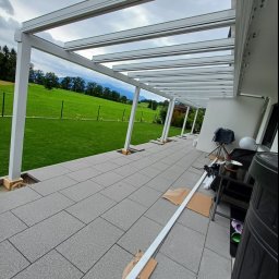 Aleksander Musioł - Montaż białej pergoli tarasowej z widokiem na zielony trawnik i góry, elementy konstrukcyjne leżą na szarych płytach chodnikowych.