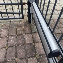 Aleksander Musioł - Automatyka otwierająca czarną, metalową bramę, widoczny siłownik z przewodami na tle kostki brukowej.