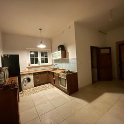 Aleksander Musioł - Ukończona aranżacja kuchni z drewnianymi szafkami, płytkami w odcieniach błękitu nad blatem, pralką w zabudowie i lampą wiszącą w stylu retro, widok na otwarty salon z drzwiami.