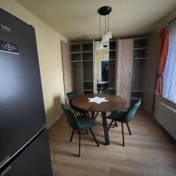 Aleksander Musioł - Jadalnia z okrągłym drewnianym stołem, zielonymi krzesłami i nowoczesną lampą wiszącą na tle szafy wnękowej i lodówki.