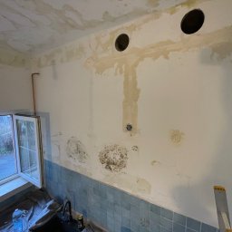 Aleksander Musioł - Ściana w łazience przed remontem z widocznymi śladami wilgoci, grzybem i przygotowanymi otworami wentylacyjnymi, niebieskie kafelki na dole ściany, okno z widokiem na zewnątrz.