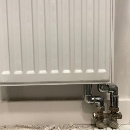 Aleksander Musioł - Biały grzejnik panelowy z chromowanymi rurami i zaworami przy podłodze w trakcie prac remontowych, widoczne zabrudzenia i resztki materiałów budowlanych.