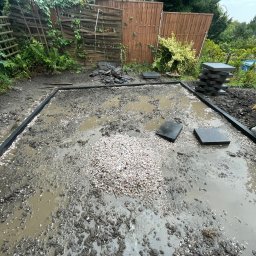 Aleksander Musioł - Przygotowanie terenu pod taras lub ścieżkę: wytyczony obrys z czarnych obrzeży, wysypany żwir, ułożone płyty chodnikowe, widoczna sterta płyt i krzesło ogrodowe w tle.