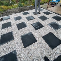 Aleksander Musioł - Układanie ścieżki z kwadratowych płyt na jasnym żwirze w ogrodzie, widoczne fragmenty roślinności w tle, osoba w szarych spodniach w trakcie pracy.